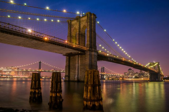 Brooklyn Bridge glowing with lights against dusk sky while river reflects warm tones. Perfect for travel, city life, architecture, and night photography themes. Suitable for wallpapers, blog posts, or marketing materials highlighting New York City's landmark views.