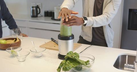 Diverse men preparing green smoothie in modern kitchen using blender and fresh greens