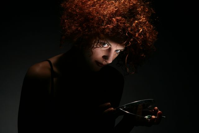 Woman with intense expression and red curly hair holding mirror in dramatic, dark lighting. Ideal for use in artistic projects, mood boards or blogs focusing on mystery, introspection or drama.