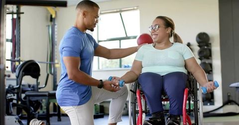 Inclusive Personal Trainer Guiding Wheelchair User Lifting Dumbbells in Bright Gym