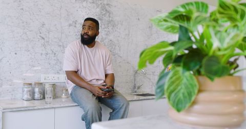 Man Relaxing in Modern Kitchen Holding Smartphone, Next to Potted Plant