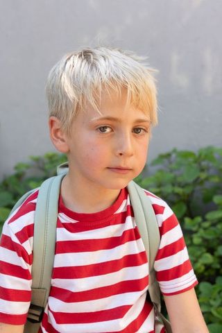 Blonde boy with backpack in garden wearing red striped shirt, back-to-school portrait