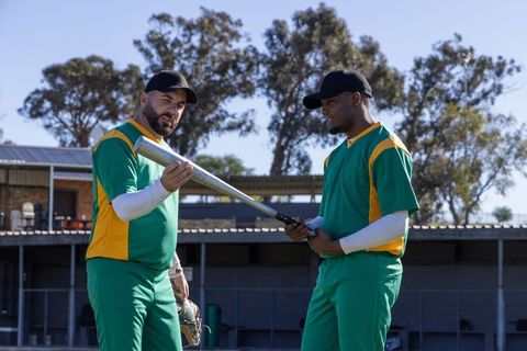 Baseball teammate coaching with bat near dugout in green and yellow uniforms