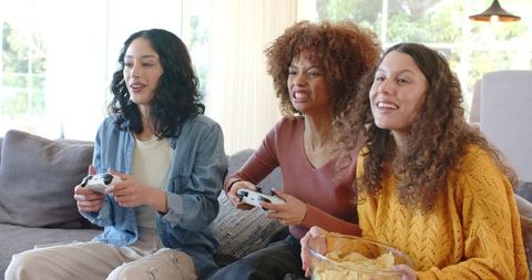 Group of friends playing video games on cozy sofa while sharing snacks and laughing