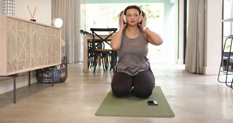 African woman kneeling on yoga mat wearing headphones using smartphone during home workout