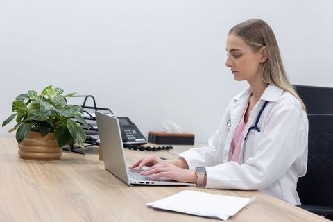 Female doctor typing on laptop at modern clinic desk with stethoscope, plant and paperwork