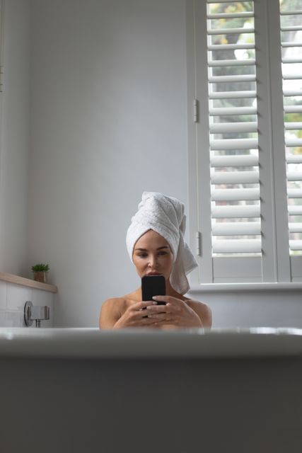Woman sitting in bathtub with towel wrapped around her head while using mobile phone, conveying relaxation and self-care. Useful for topics on modern lifestyle, home bath routines, or the intersection of technology and daily life.
