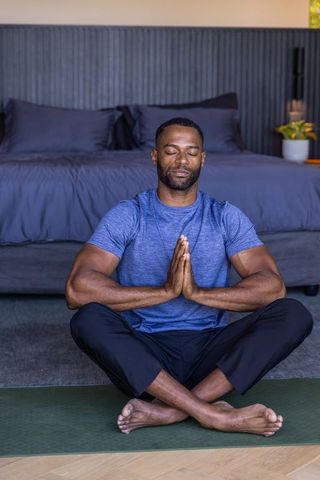 Man meditating cross-legged on green yoga mat inside minimalist bedroom for wellness