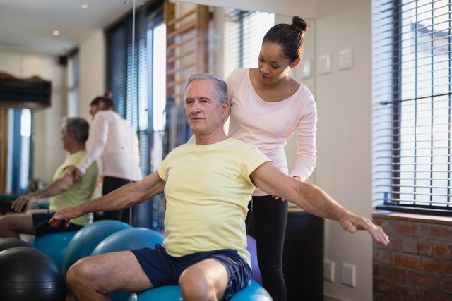 Female therapist assisting senior male patient during a physical therapy session. The patient is seated on an exercise ball with arms outstretched, focusing on rehabilitation and recovery. Ideal for use in healthcare, physiotherapy, elderly care, and wellness content.