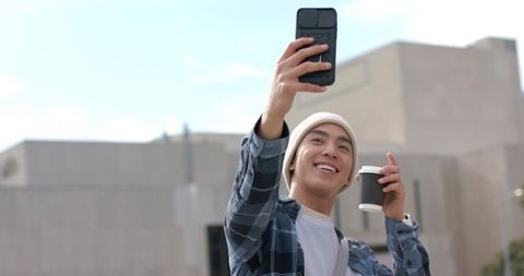 Young Asian man taking selfie with takeaway coffee on urban campus smiling casually