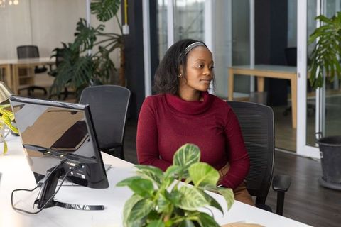 African american woman sitting at coworking desk with greenery using monitor