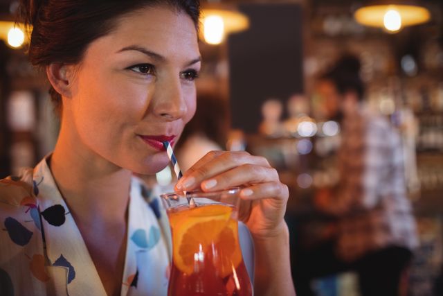 Young woman sipping a cocktail with an orange slice garnish through a striped straw in a pub. Ideal for use in advertisements for bars, pubs, or restaurants, as well as lifestyle blogs and social media posts about nightlife, leisure, and socializing.