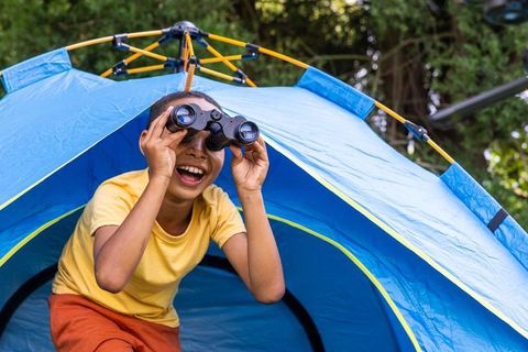 African boy using binoculars at blue pop-up tent laughing during backyard camping fun