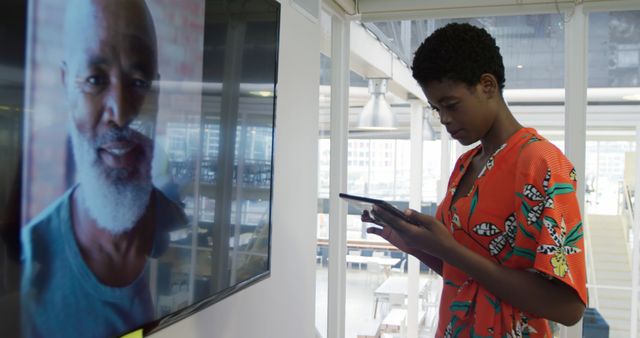 Young African woman stands in a modern office using a tablet device while engaging in a video call. This can be used in contexts related to workplace technology, remote communication, modern work environments, or tech-savvy professionals.