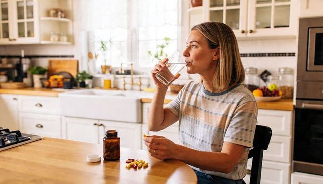 Woman drinking water and taking supplements at sunlit farmhouse kitchen table for wellness