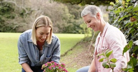 Women tending pink potted flowers kneeling in backyard garden smiling together gardening