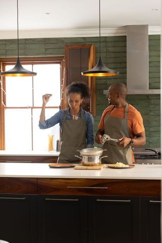 Couple cooking together on kitchen island sprinkling seasoning stirring pot in aprons