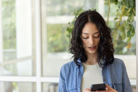 Indian woman using smartphone in sunlit minimalist workspace wearing denim shirt
