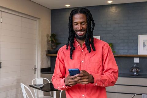 Smiling african american man texting on smartphone in cozy modern kitchen wearing coral shirt