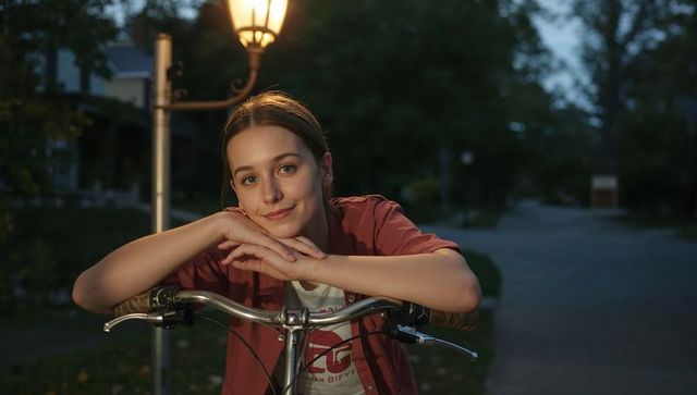 Young woman leaning on bicycle handlebars under streetlamp at dusk in peaceful suburb