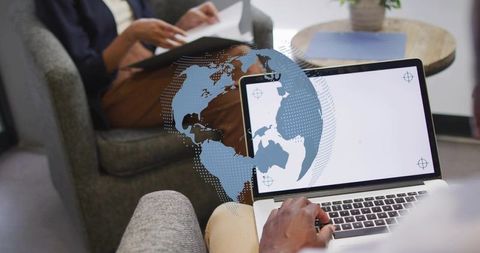 Typing on laptop showing global overlay while colleague reading papers in lounge