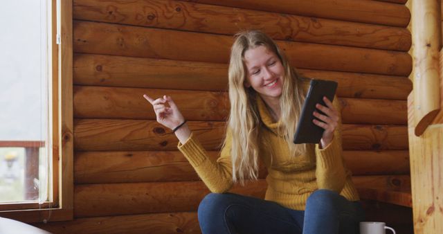 Smiling woman in casual clothing enjoying video chat on tablet in cozy wooden cabin. Useful for promoting remote work, digital communication, leisure activities, and travel destinations.