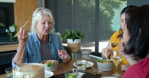 Mature women sharing noodle bowls using chopsticks around modern home dining table