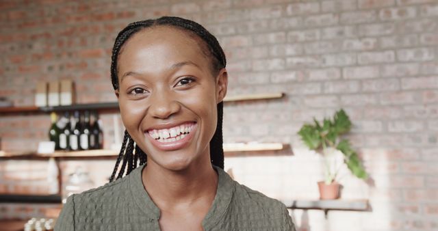 The image features a young woman with braided hair smiling brightly in a cozy indoor setting. The background includes a brick wall, shelves with bottles, and a potted plant, indicating a warm, welcoming environment. This image is perfect for marketing materials related to positive experiences, lifestyle blogs, advertisements promoting inclusivity and happiness, or corporate headshots promoting a modern and approachable atmosphere.