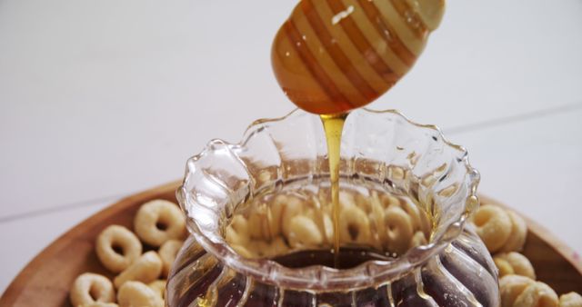 Honey dipper drizzling honey into glass bowl surrounded with small round doughnuts. Ideal for promoting natural sweeteners, healthy breakfast options, or food and beverage presentations. It can be used in blogs, cooking websites, healthy lifestyle marketing, or recipe articles.