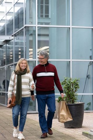 Mature couple walking urban plaza holding shopping bag and smartphone casual outing
