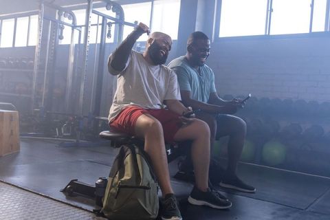 Two african men relaxing on gym bench using smartphones after workout showing camaraderie
