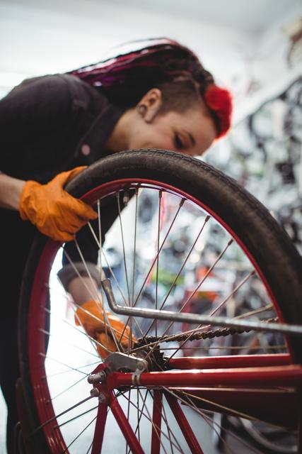 This image captures a female mechanic skillfully repairing a bicycle tire in a workshop. Wearing orange gloves, she appears deeply focused on her task. The background suggests a busy repair shop. This photo can be used to depict concepts like mechanical skills, industrial professions, women's empowerment in trades, or promotional material for bike repair services.