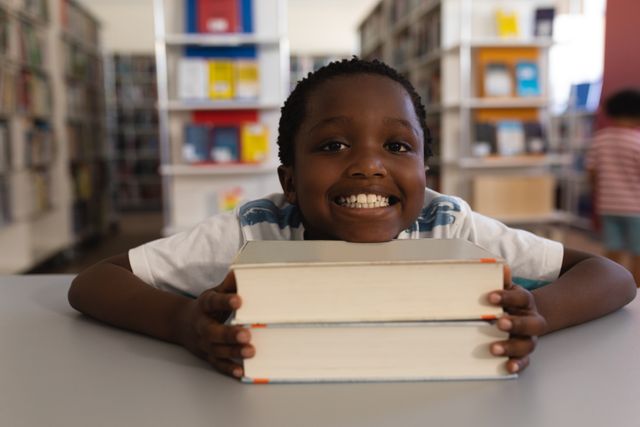 Front view of happy schoolboy leaning his face on books and looking at camera on table in school library