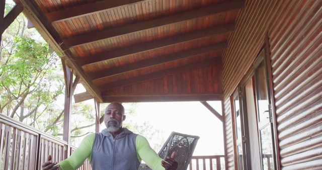 Mature man practicing meditation on a wooden cabin deck. He is sitting cross-legged, looking peaceful and serene with trees visible in the background. Ideal for concepts of mindfulness, relaxation, finding inner peace, and outdoor well-being. Perfect for wellness blogs, meditation apps, lifestyle articles, and nature retreats.