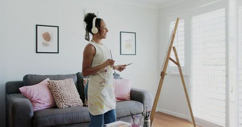 African american woman painting in bright home studio wearing headphones by easel
