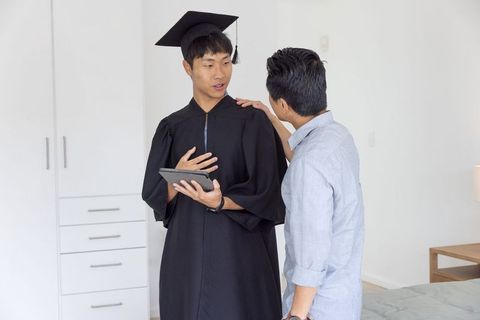 Asian graduate celebrating with father at home holding tablet, wearing cap and gown