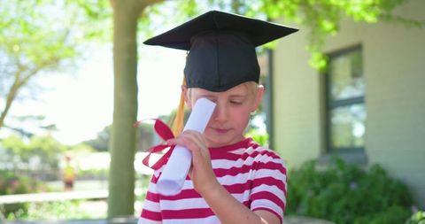 Young boy wearing graduation cap holding diploma scroll outdoors on school campus