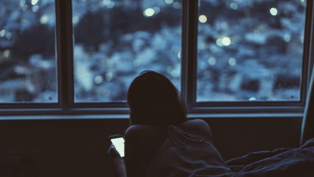 Person lying near a window, illuminated by the soft glow of a smartphone screen, overlooking city lights. Ideal for depicting modern urban lifestyle, technology use, personal downtime, evening rituals, and moments of solitude.