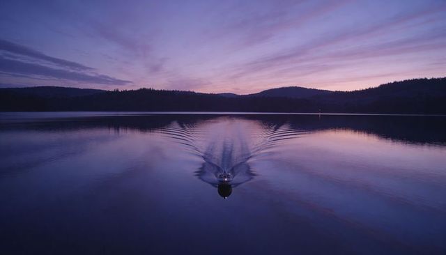 Small motorboat gliding across tranquil lake at dusk leaving v-shaped wake under pastel sky