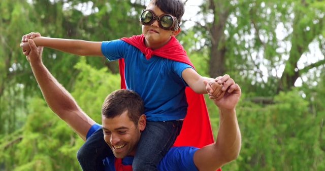 A child dressed as a superhero, complete with cape and goggles, enjoys a piggyback ride on his father's shoulders. Both are outside in a lush green park, embodying the spirit of adventure and bonding. Ideal for illustrating family activities, the importance of imagination in childhood, or outdoor recreational campaigns.