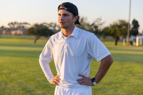 Young athlete standing hands-on-hips on sunlit grass field wearing white kit and backward cap