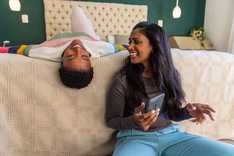 Multicultural mother and teen daughter laughing in bedroom with daughter lying upside down