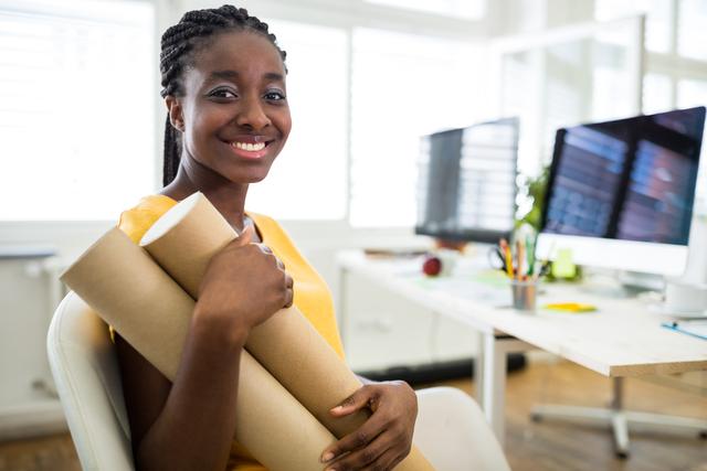 Young African American female graphic designer smiling while holding a chart holder in a modern office. Ideal for use in business, professional, and creative industry contexts, showcasing diversity and modern work environments.