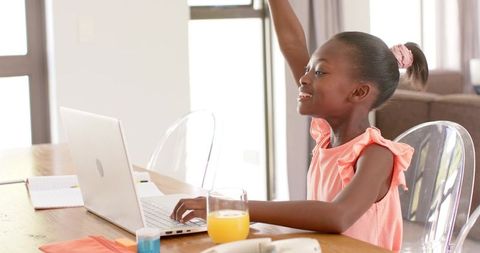 African american girl raising hand while learning online at dining table with laptop