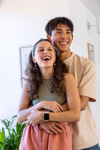 Asian and african american couple hugging and laughing in sunlit home with smartwatch