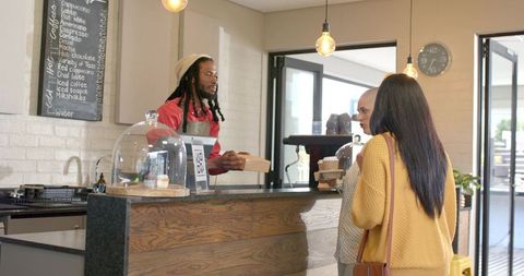 Barista handing pastry box to diverse customers at cozy wooden cafe counter with chalkboard