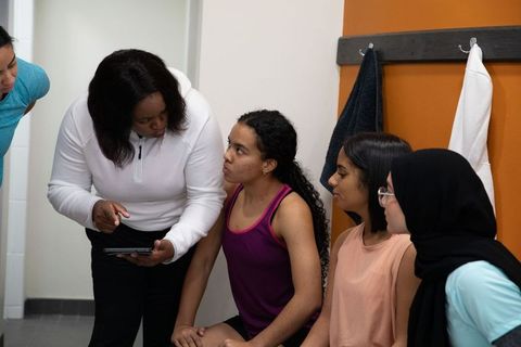 African american coach mentoring diverse teen girls in locker room reviewing tablet