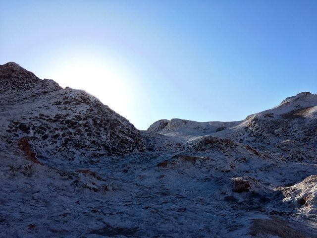 Snow-covered rocky hills illuminated by sunlight under a clear blue sky evoke a serene, desolate winter landscape. Ideal for use in travel websites, nature blogs, environmental articles, or seasonal advertising campaigns to illustrate winter beauty and natural landscapes.