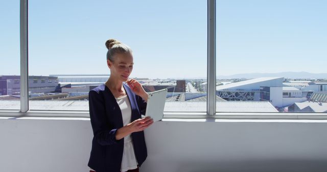 This photo shows a professional woman standing by a window holding a digital tablet. The cityscape outside the large window indicates an office setting. Ideal for use in business, technology, entrepreneurship, and modern workplace themes in articles, presentations, and advertisements.