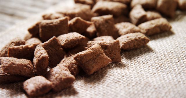 Chocolate-flavored cereal pieces are scattered across a textured beige cloth, creating a tempting and delicious-looking scene. Perfect for a quick breakfast or a sweet snack, these bite-sized treats appeal to anyone with a love for chocolatey goodness.